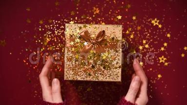 女人手打开礼盒。 闪耀着金色的星星，闪闪发光的纸屑。 在红色背景下打开包装礼物